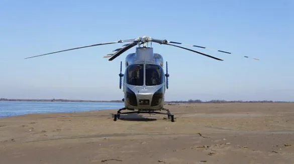 A silver helicopter with its rotors spread, parked on a sandy riverbank under a clear blue sky.