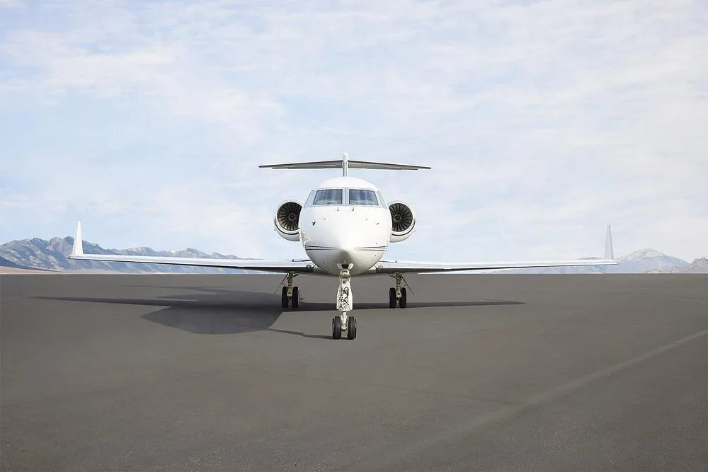Front view of a white Gulfstream G550 business jet on a tarmac with mountains in the distance.
