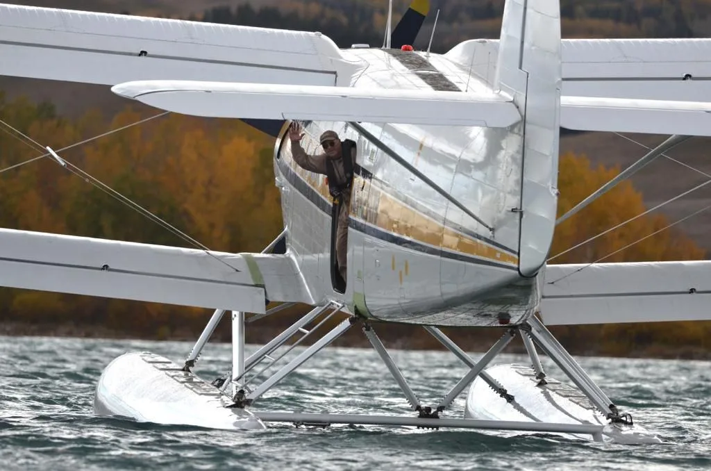 A person waves from the open door at the rear of a white and yellow Antonov AN-2 floatplane on a lake.
