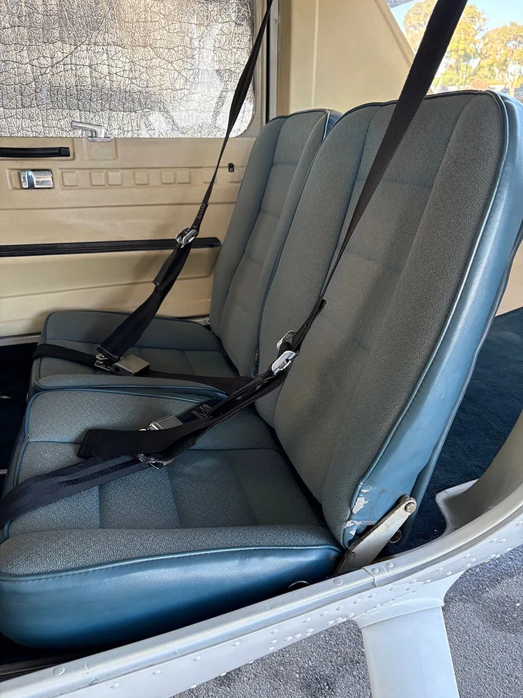Two blue fabric and vinyl aircraft seats with black seatbelts in the rear cabin, with a window sunshade visible.