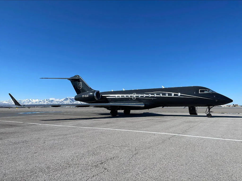 A black business jet with white stripes and registration TT-CLG parked on an airfield with snow-capped mountains in the background under a clear blue sky.