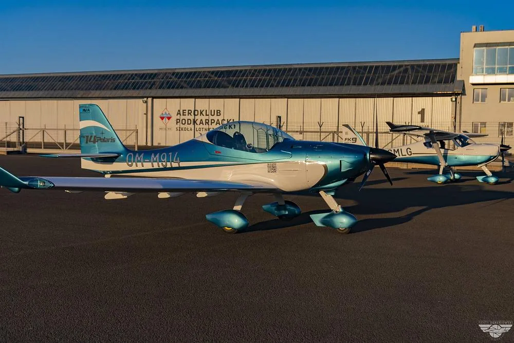 A blue and white FK14 Polaris aircraft (OM-M914) parked on an airfield in front of a hangar, with another aircraft behind it.