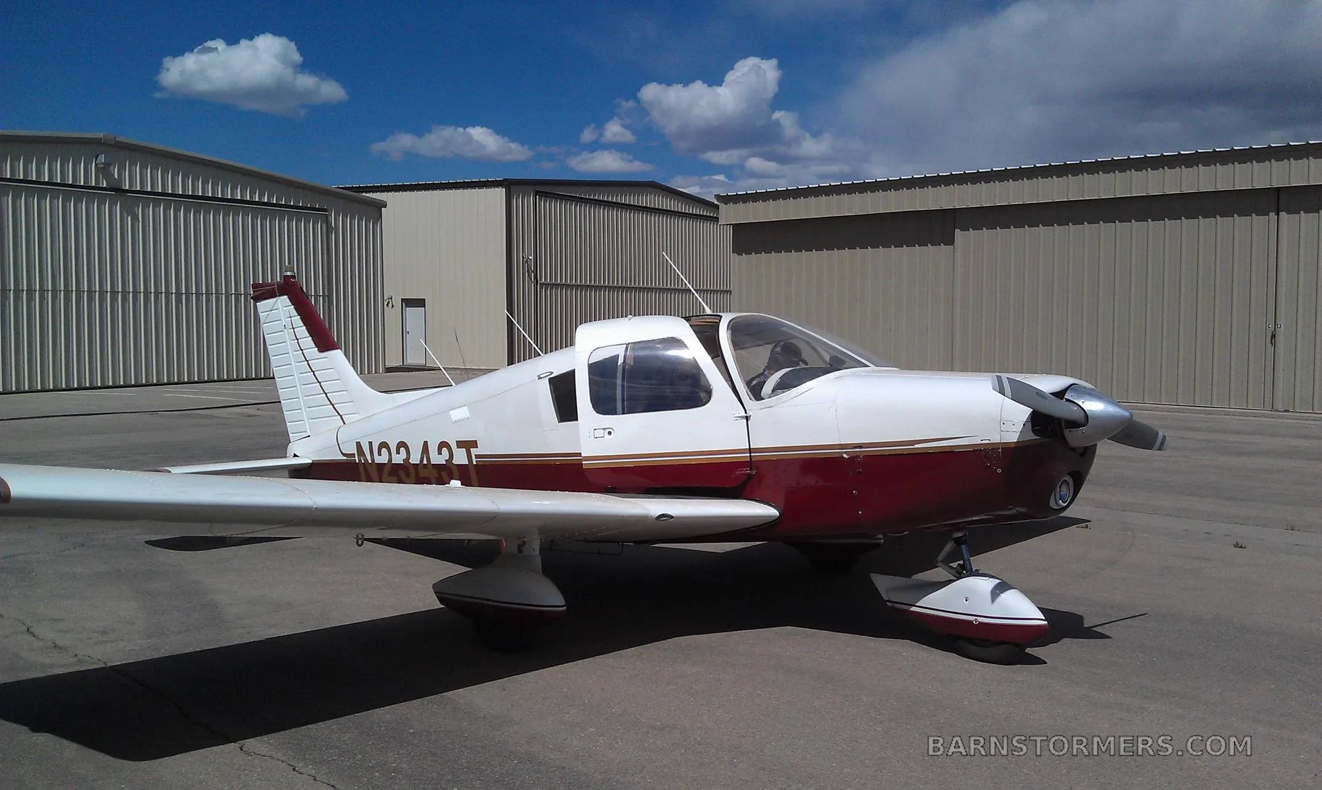 A white and maroon Piper Cherokee 140 (N9243T) parked on a tarmac in front of hangars under a blue sky.