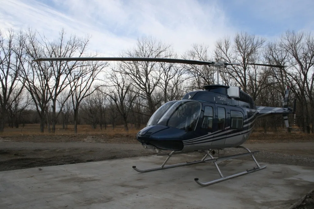 Front-side view of the blue and white Bell 206L helicopter (N60VG) parked on a concrete pad with bare trees in the background.