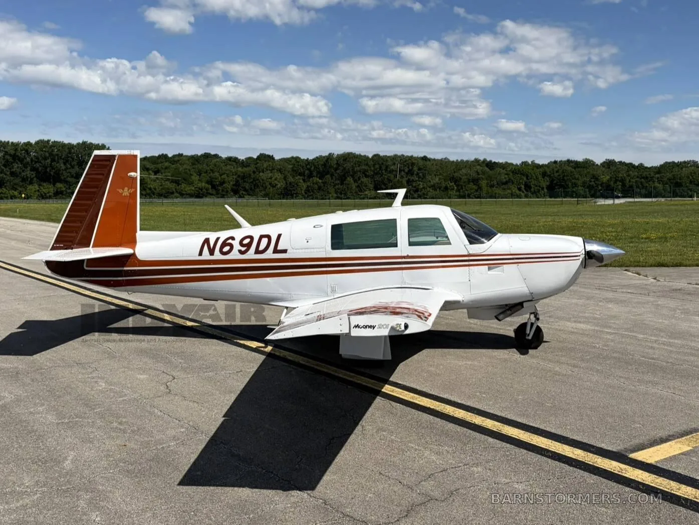 A white and brown Mooney M20J aircraft (N69DL) is parked on an asphalt runway under a partly cloudy sky, viewed from the right side.