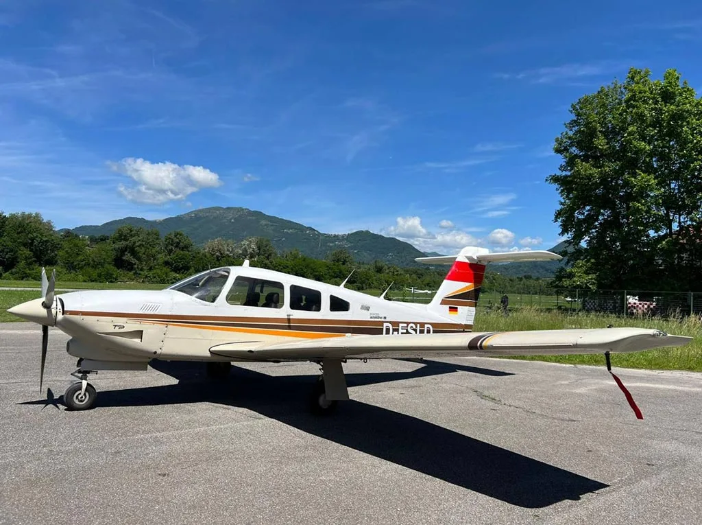 A white and brown Piper Arrow III (D-ESLD) parked on an asphalt apron with mountains in the background under a blue sky.