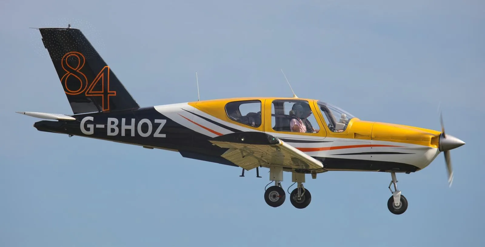 A yellow, black, and white propeller aircraft with registration G-BHOZ and number 84 on the tail, flying with landing gear extended.