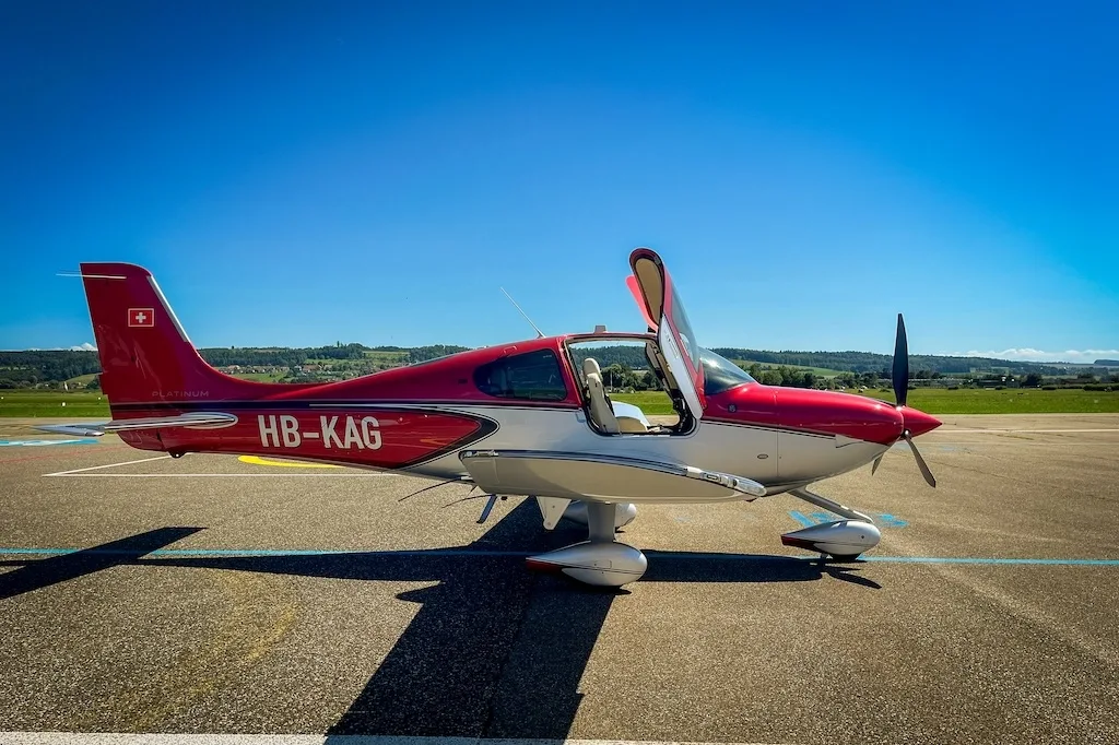 Side view of a red and silver Cirrus SR22 aircraft with doors open, parked on an airfield under a clear blue sky.