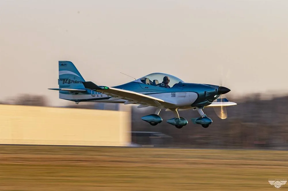 A blue and white FK14 Polaris aircraft (OM-M914) with a pilot in the cockpit, flying low over a grassy field.