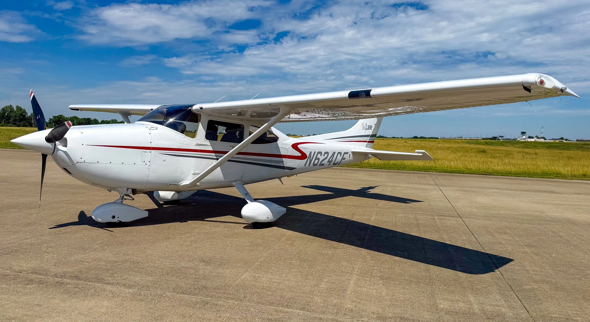 A white Cessna 182 Skylane with red and gray stripes, registration N624CF, parked on an asphalt ramp under a partly cloudy blue sky.