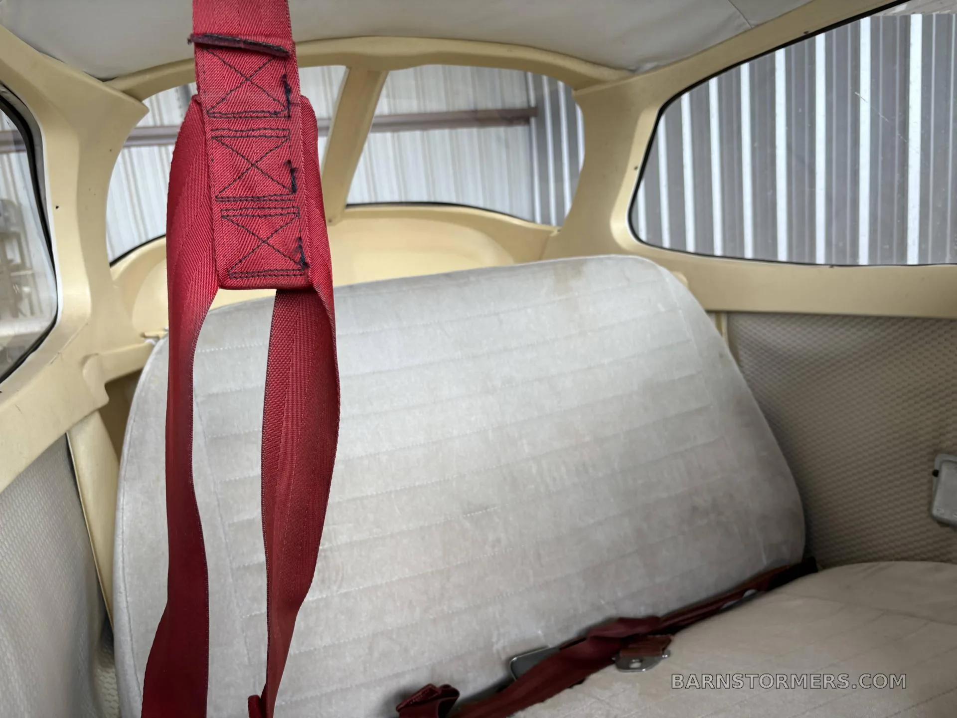 The rear interior of a small aircraft cabin, showing a light-colored bench seat and a red shoulder harness.