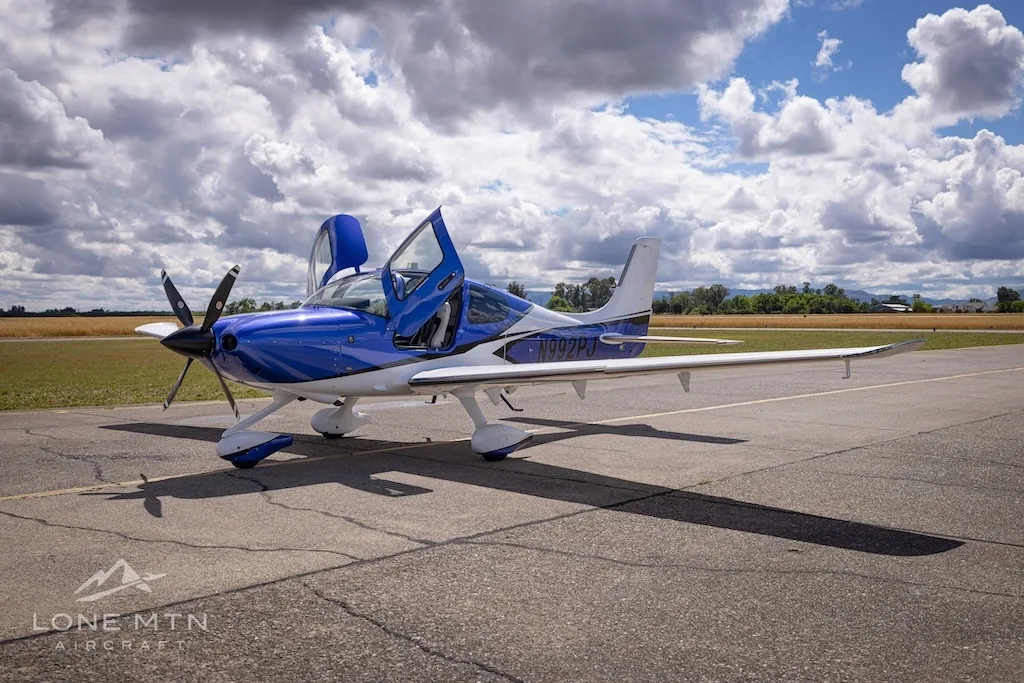A blue and white Cirrus SR22 aircraft with its doors open, parked on an airfield under a cloudy sky.