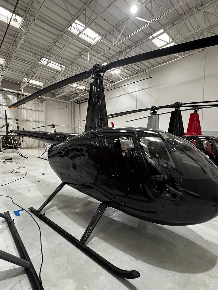 A black Robinson R44 helicopter is parked in a hangar, viewed from the front-right.