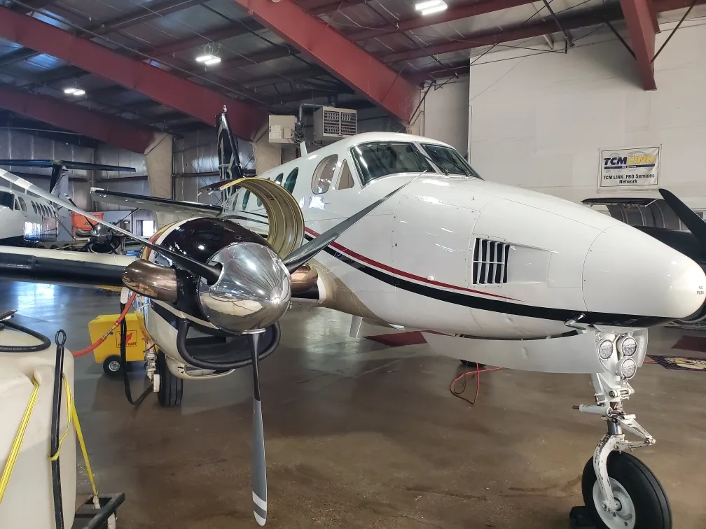 A white Beechcraft King Air 90 turboprop aircraft with black and red stripes on the fuselage, parked in a hangar, viewed from the front-right.
