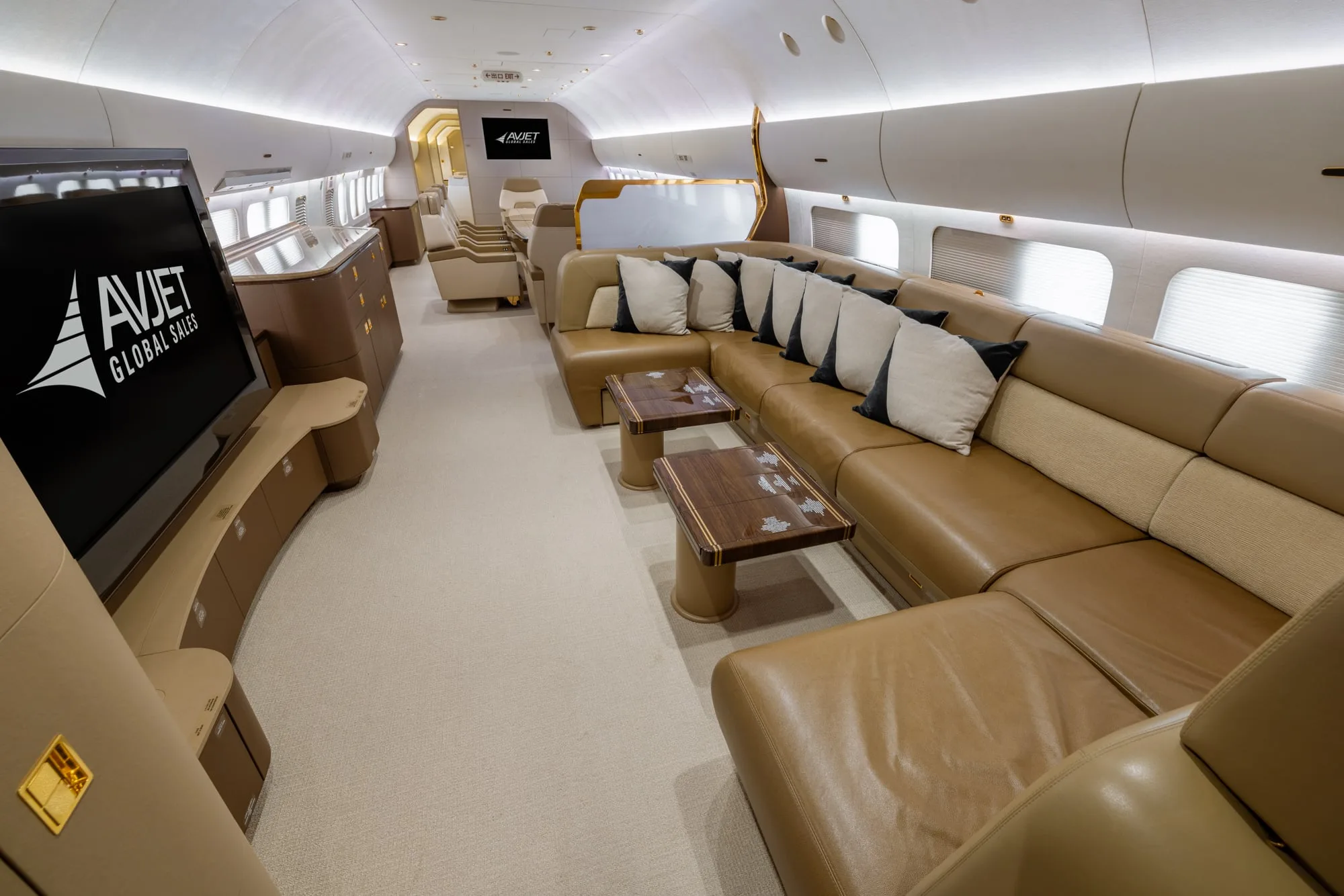 A spacious private jet cabin interior with a large brown leather sectional sofa, coffee tables, and a large screen displaying the Avjet logo.