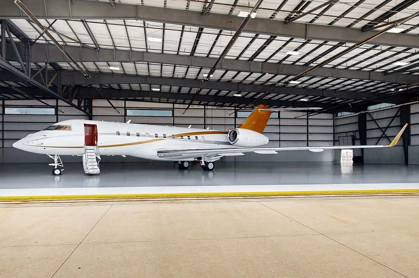 A white business jet with gold accents on the tail and fuselage is parked in a hangar with its main door open and stairs extended.