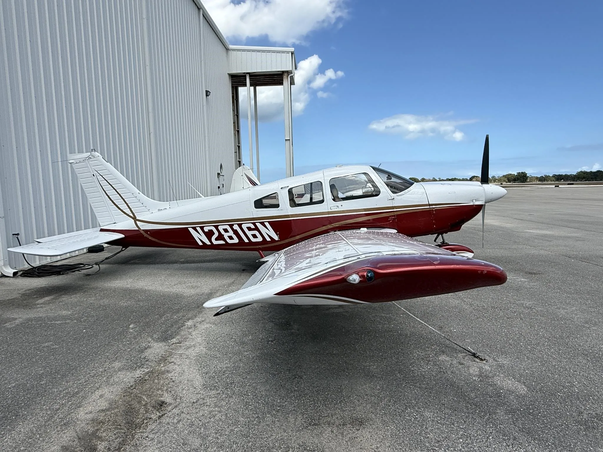 A front-side view of a white and red Piper PA-28-161 aircraft, N2816N, parked on an asphalt ramp next to a white hangar.