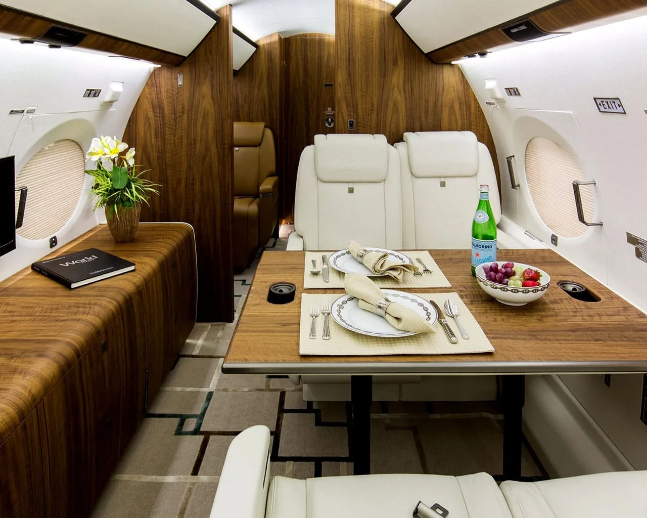 A private jet cabin dining area with a set table for two, white leather seats, and dark wood accents.