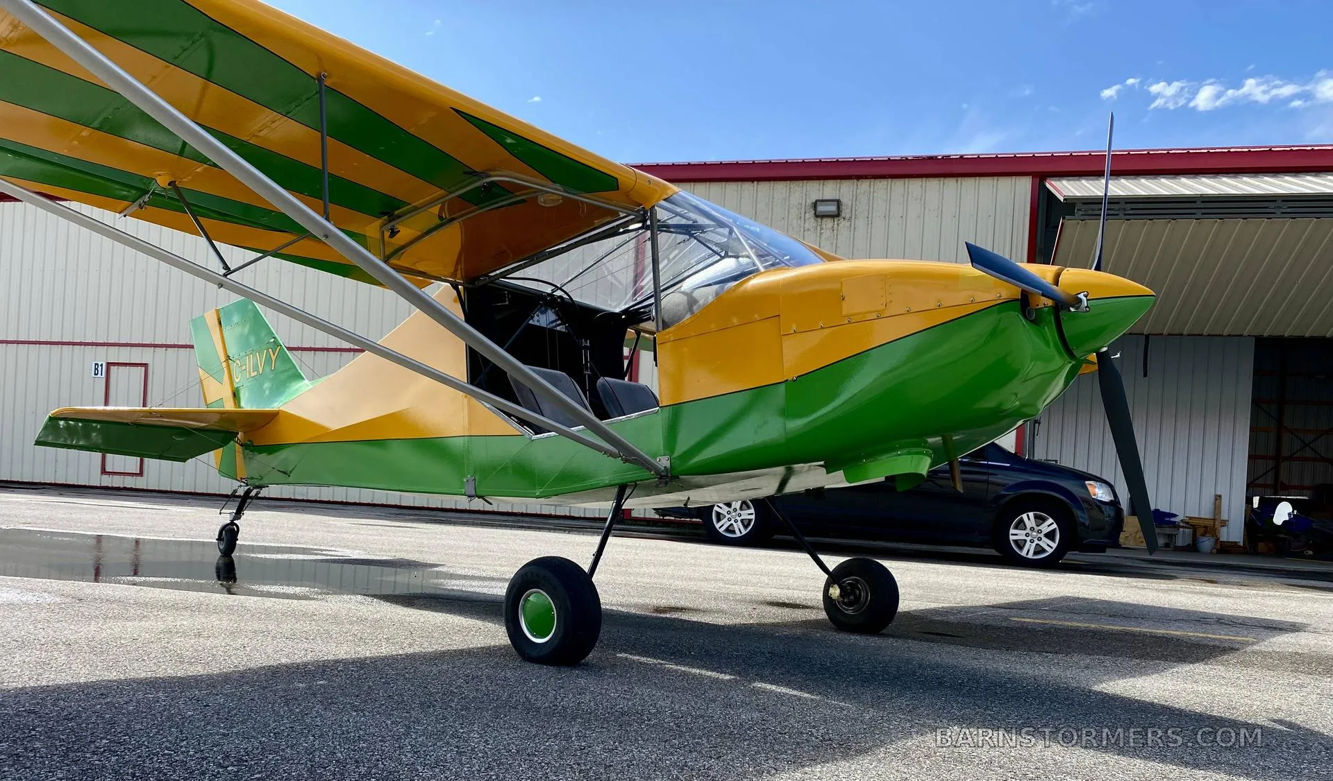 A close-up side view of a yellow and green high-wing aircraft with its door open, parked on an airfield next to a building.