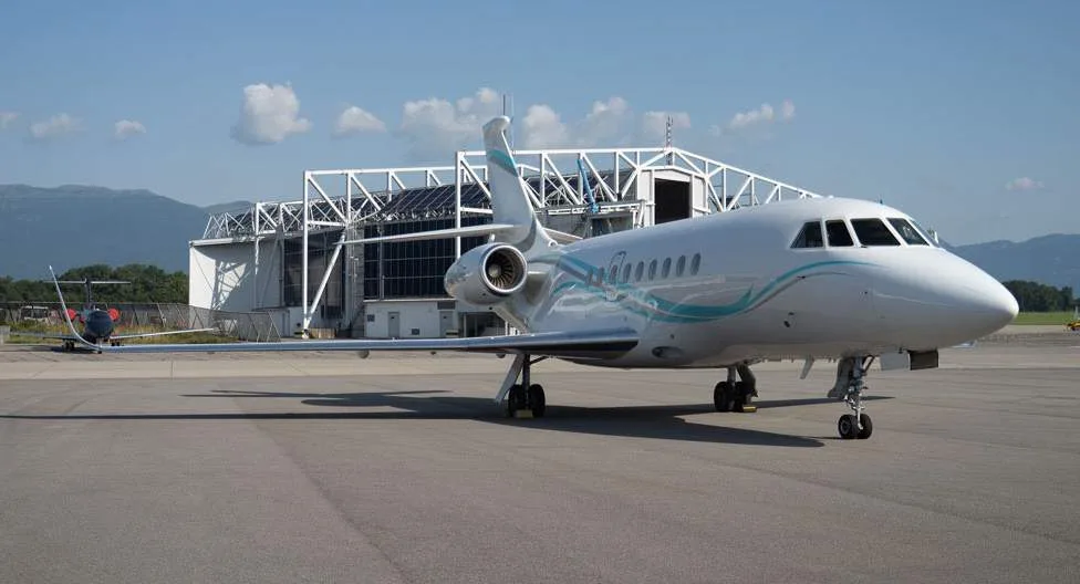 A white business jet with blue and teal stripes, parked on an airfield with a large hangar and mountains in the background.