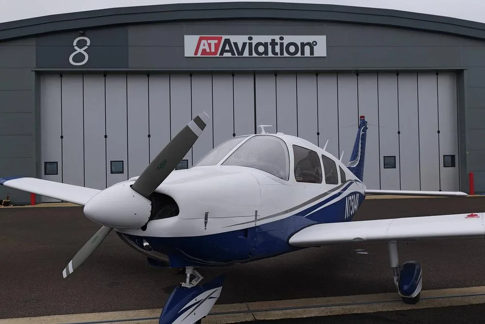 A white and blue single-engine aircraft, likely a Piper Cherokee, parked closely in front of an AT Aviation hangar.