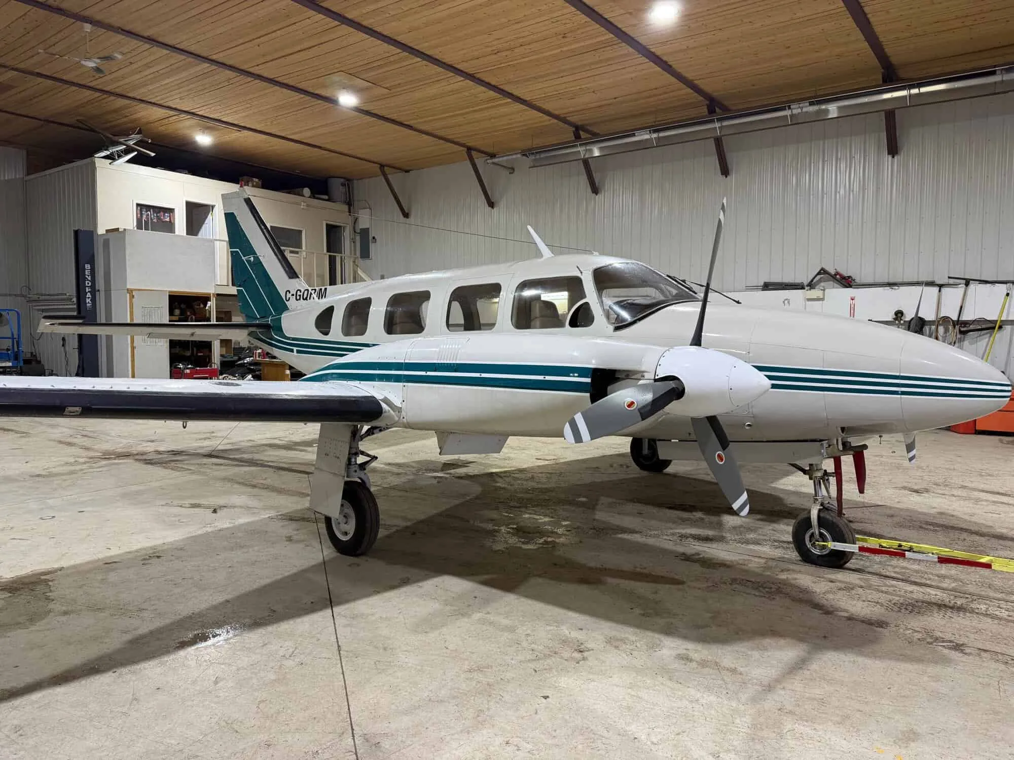 A white twin-engine Piper aircraft with green stripes and registration C-GQRIM is parked inside a hangar, viewed from the front-right.