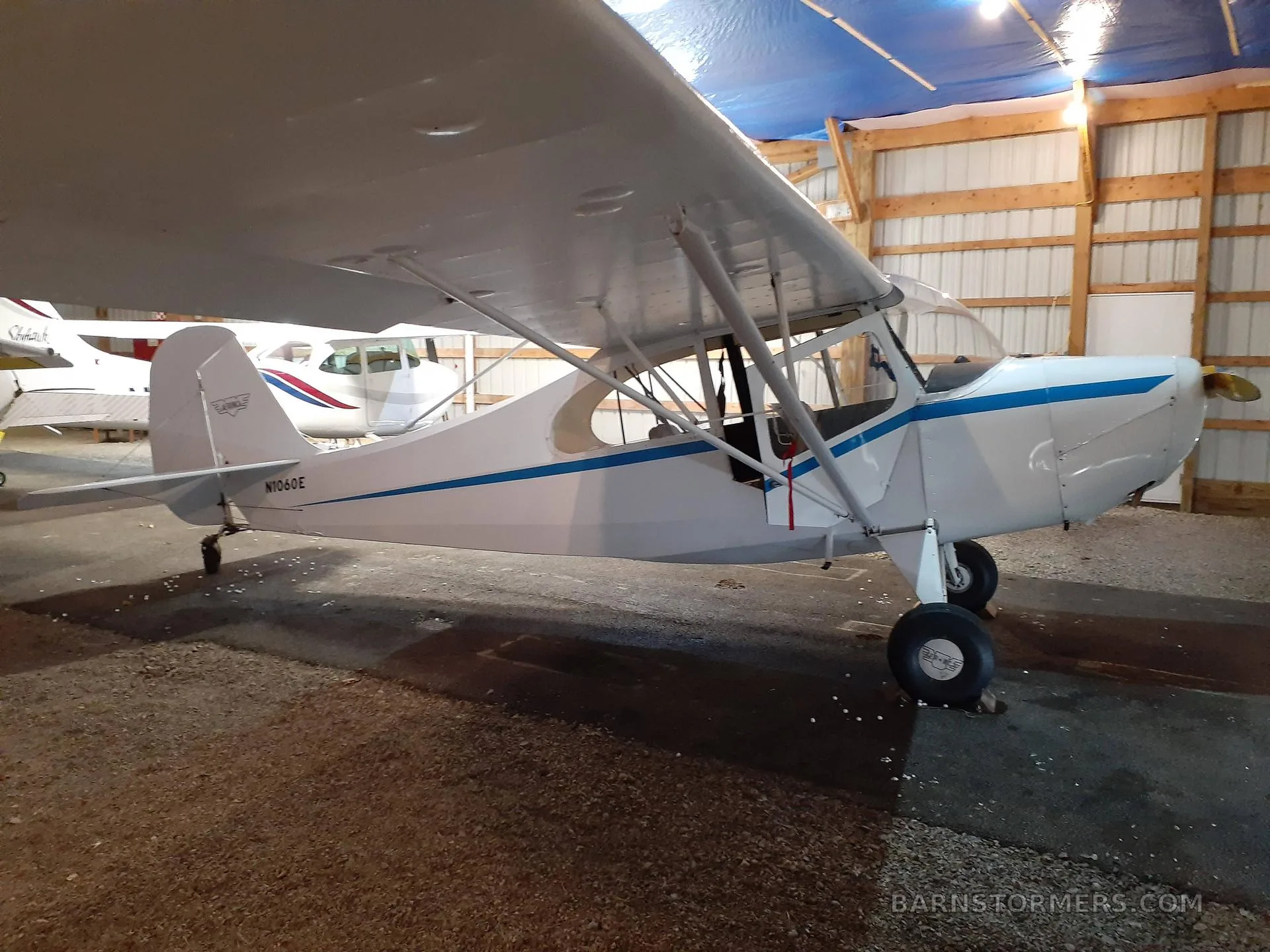 A white high-wing taildragger aircraft with blue stripes and tail number N1060E is parked in a hangar, with another aircraft partially visible in the background.
