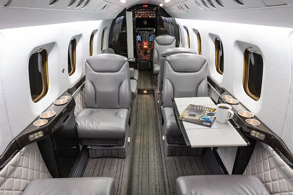 The luxurious gray leather interior of a business jet cabin with a table, magazines, and a coffee cup.