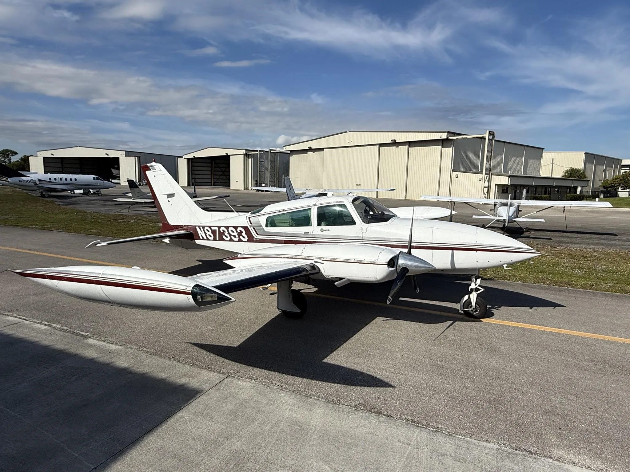A white twin-engine propeller aircraft with maroon stripes, registration N87393, parked on an asphalt ramp with hangars in the background under a partly cloudy sky.