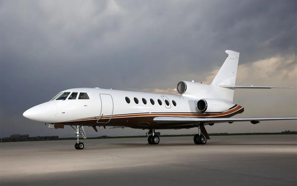 A white business jet with brown and gold stripes is parked on an airfield under a cloudy sky.
