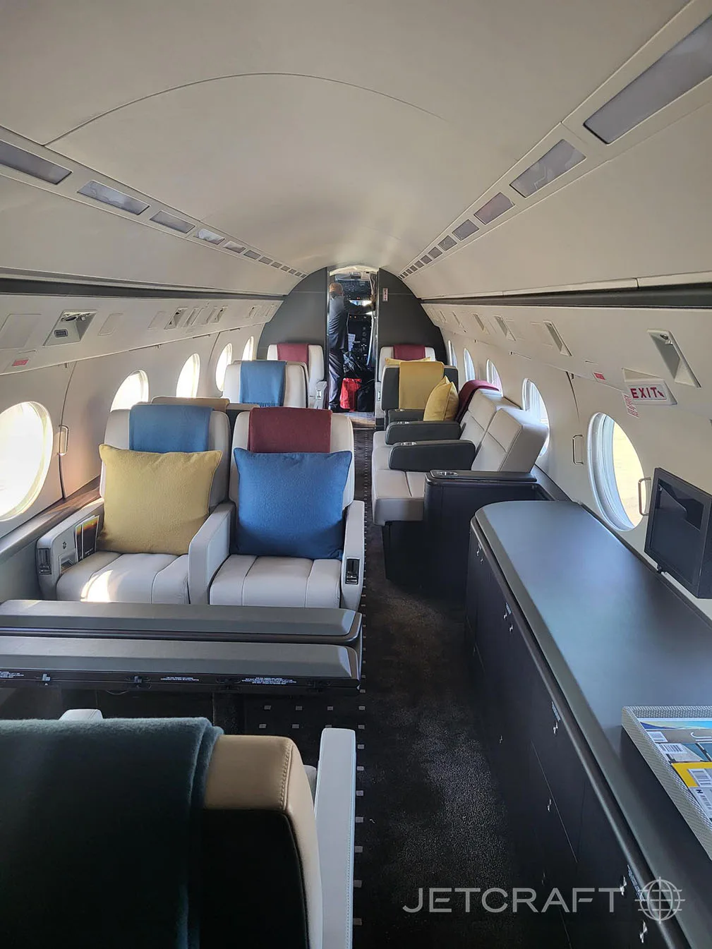 The forward section of a business jet cabin with light grey seats, colorful pillows, and a dark credenza on the right.