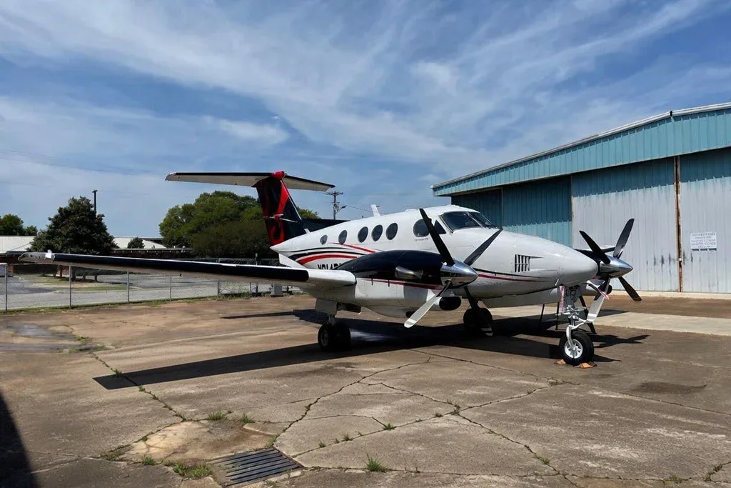A white and black Beechcraft King Air with red accents on the tail, parked on a concrete pad next to a light blue hangar under a partly cloudy sky.