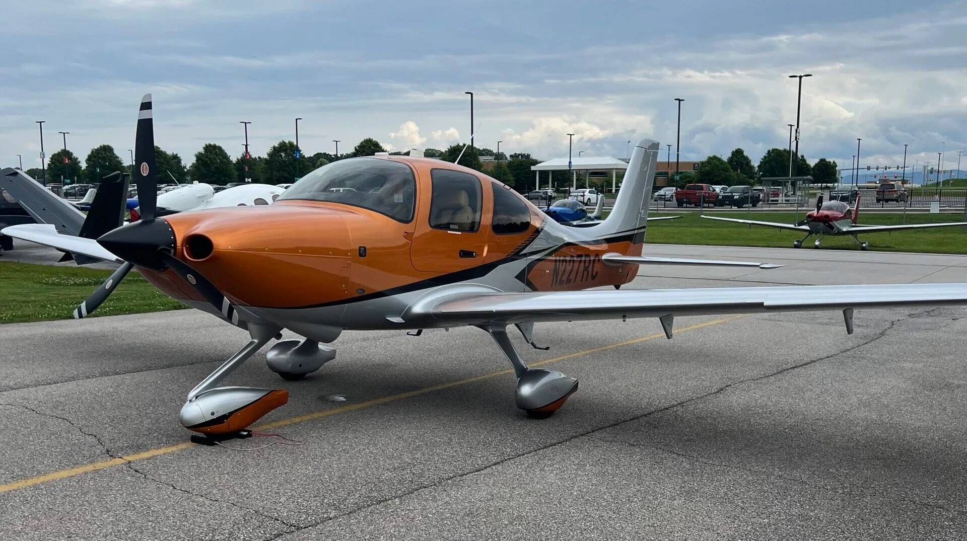 A Cirrus SR22 aircraft with an orange and silver paint scheme parked on an airfield under a cloudy sky.