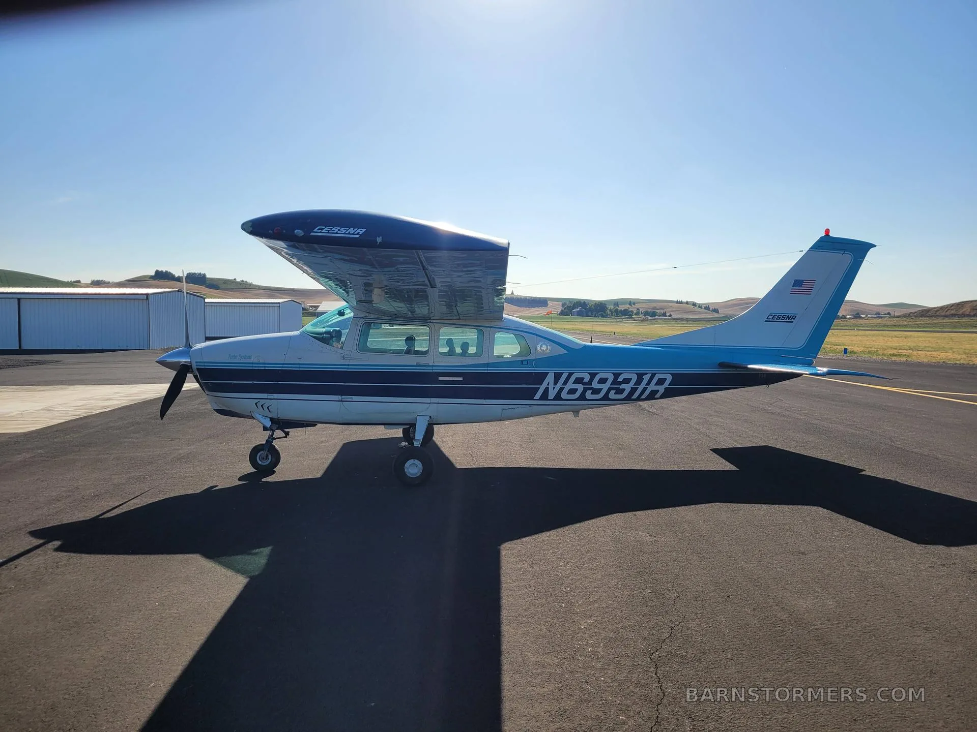 A side view of a blue and white Cessna T206H Turbo Stationair (N6931R) parked on an airfield under a bright sky.
