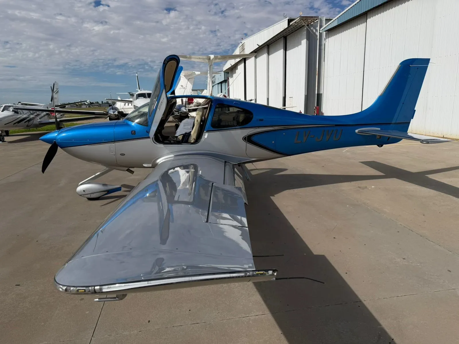 A blue and silver Cirrus SR22 aircraft, registration LV-JYU, with its left door open, parked on an airfield.