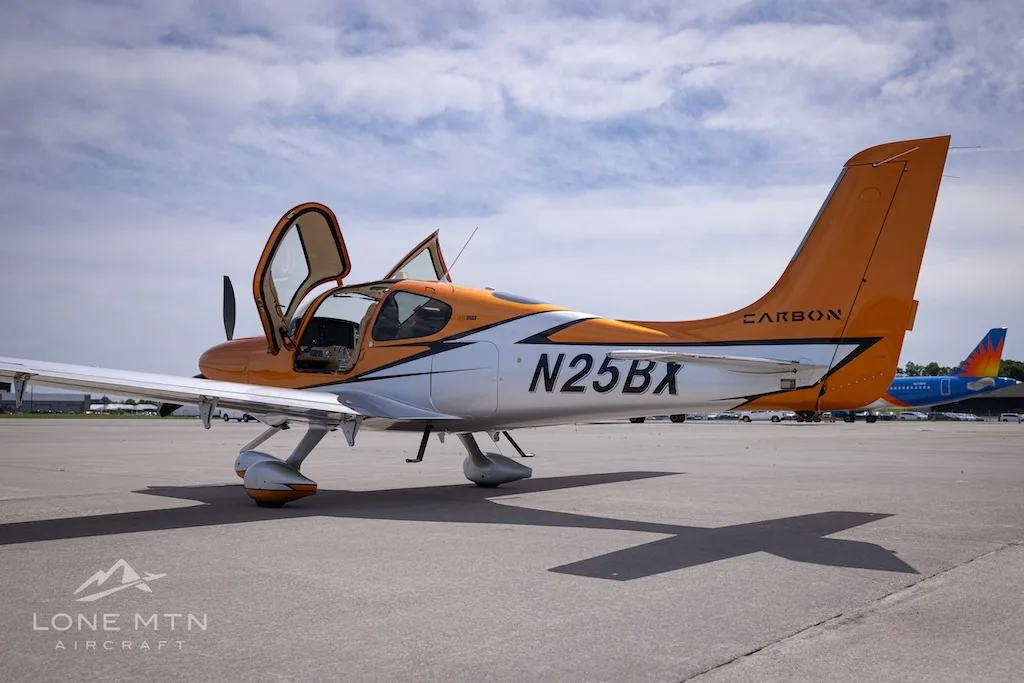 A rear-left view of the orange and silver Cirrus SR22T N25BX with its doors open on an airfield.