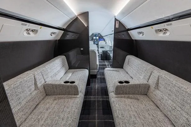 A private jet cabin section with two facing grey sofas, each with cup holders, and a dark plaid carpet.