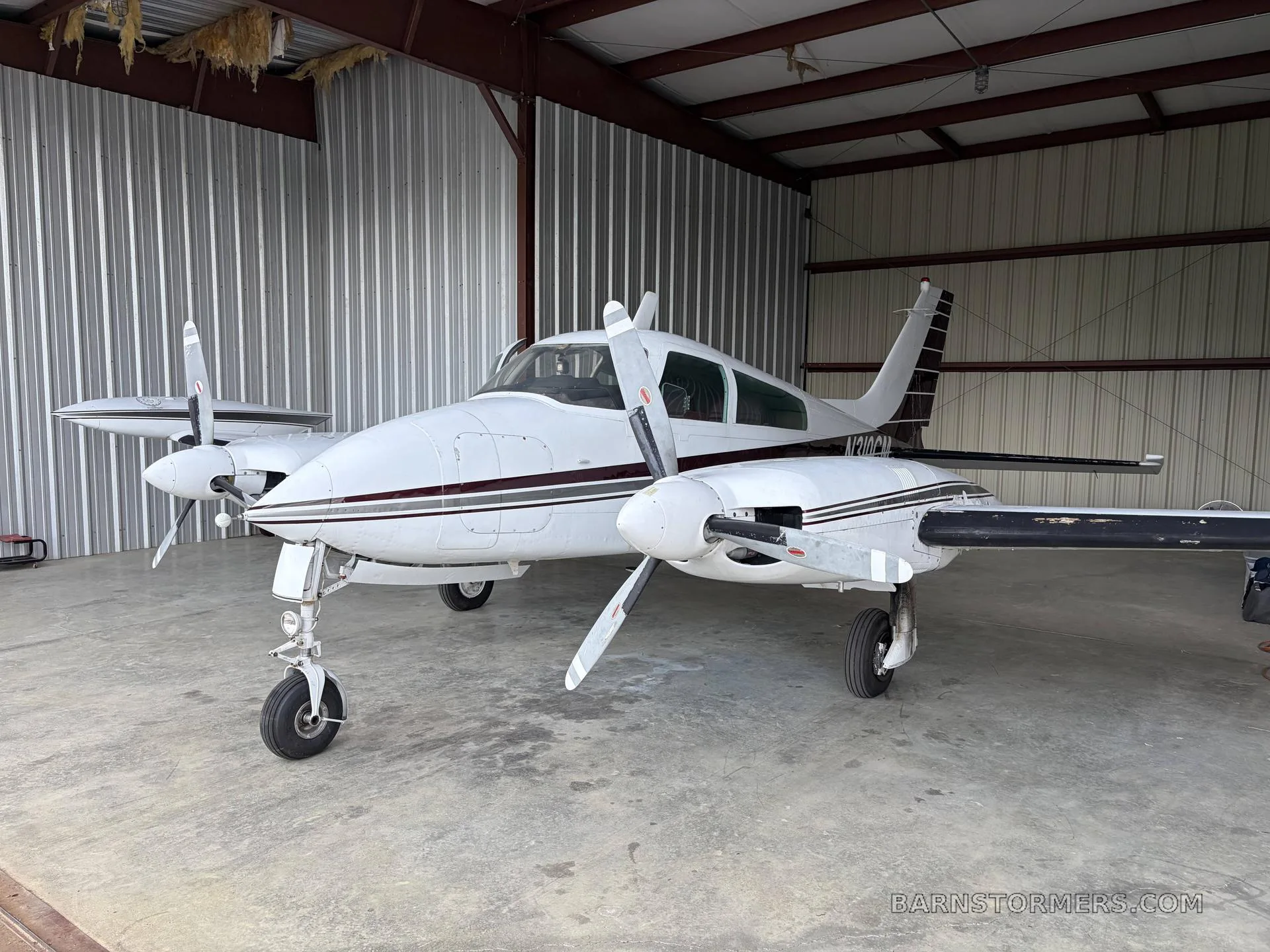 A white twin-engine propeller aircraft with dark stripes is parked inside a hangar, viewed from the front-left.