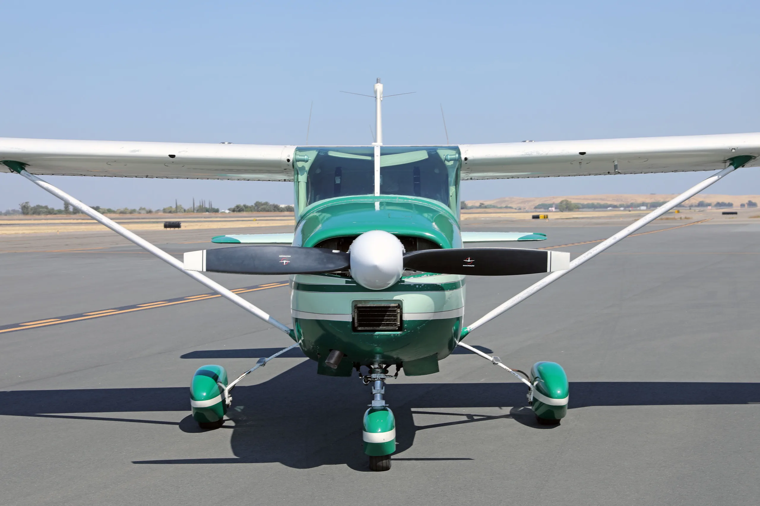 A head-on view of a green and white Cessna 182 Skylane with its propeller centered on an airfield.