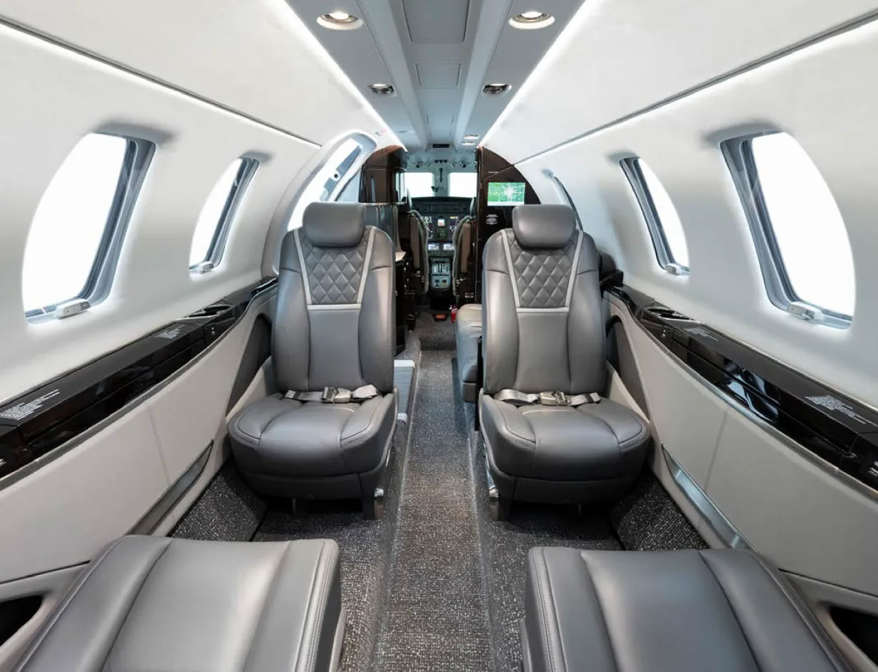 A view down the aisle of a business jet cabin, showing two rows of grey leather seats and the cockpit in the distance.