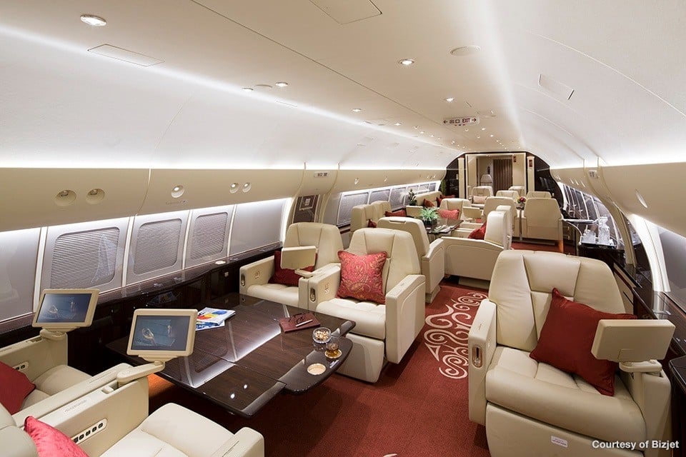 A luxurious aircraft cabin interior with cream leather seats, red pillows, and a dark wood table.