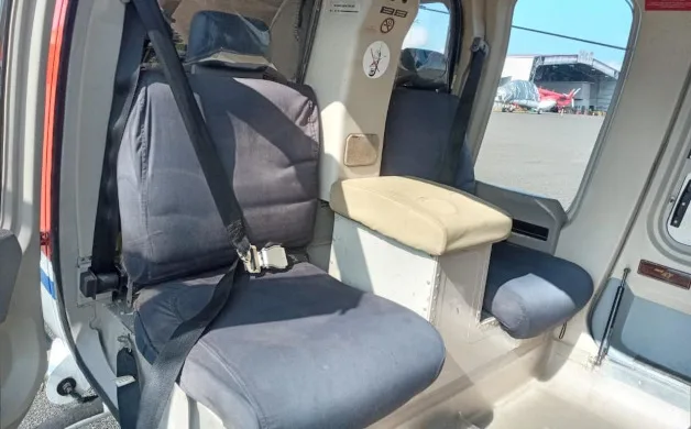 A close-up view of the dark grey passenger seats with seatbelts inside a helicopter cabin, with an aircraft visible in the background.