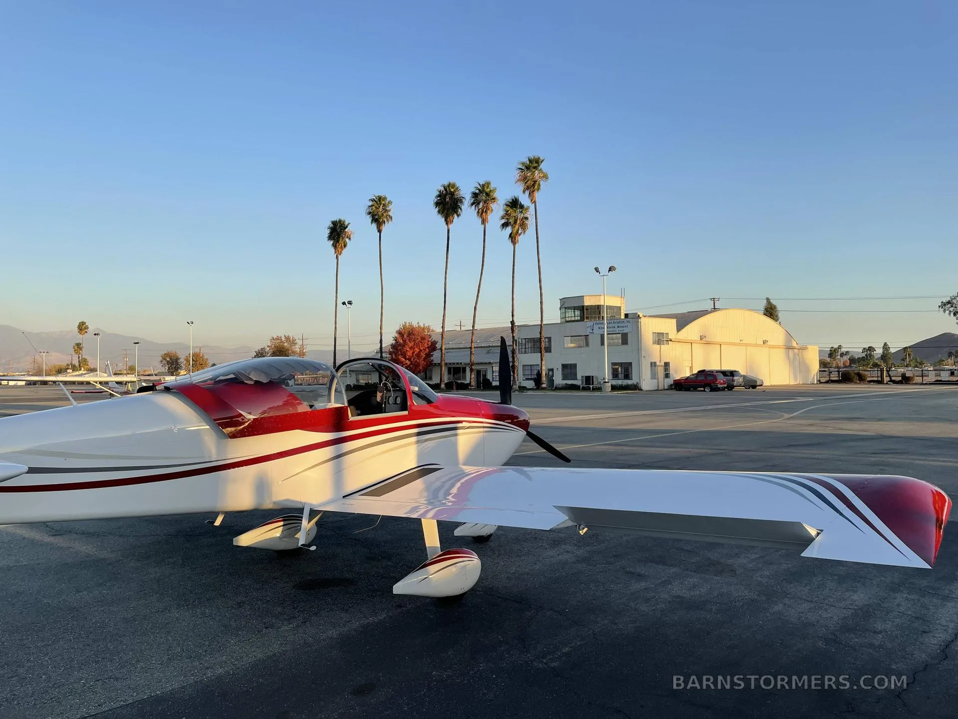 A red and white single-engine aircraft (N34BE) is parked on an airfield at sunset, with palm trees and hangars in the background.