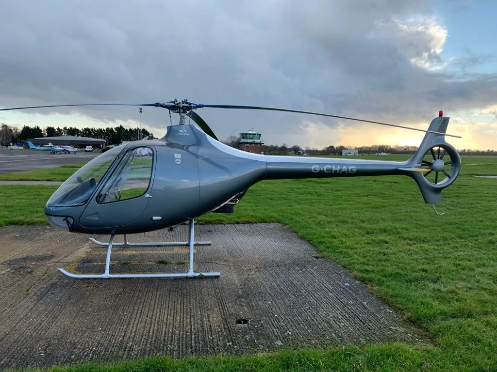 A grey Guimbal Cabri G2 helicopter with registration G-CHAG parked on a tarmac at an airfield under a cloudy sky.