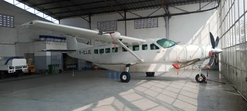 A white Cessna 208 Caravan aircraft with registration F-OJJE is parked inside a hangar, viewed from the front left.