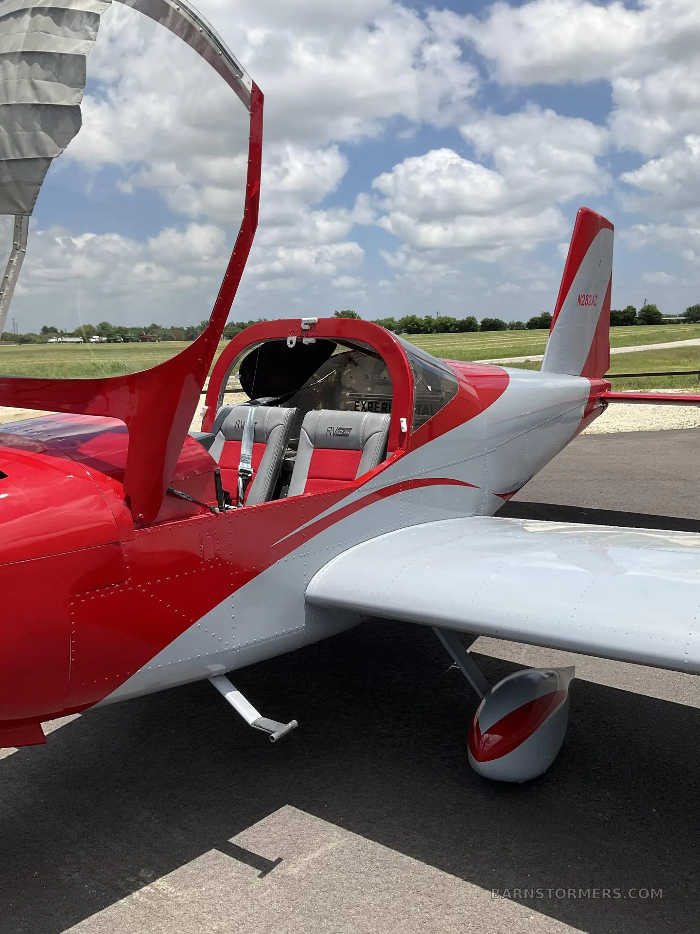 A red and grey single-engine aircraft with its canopy open, revealing two grey and red seats inside, parked on an airfield.