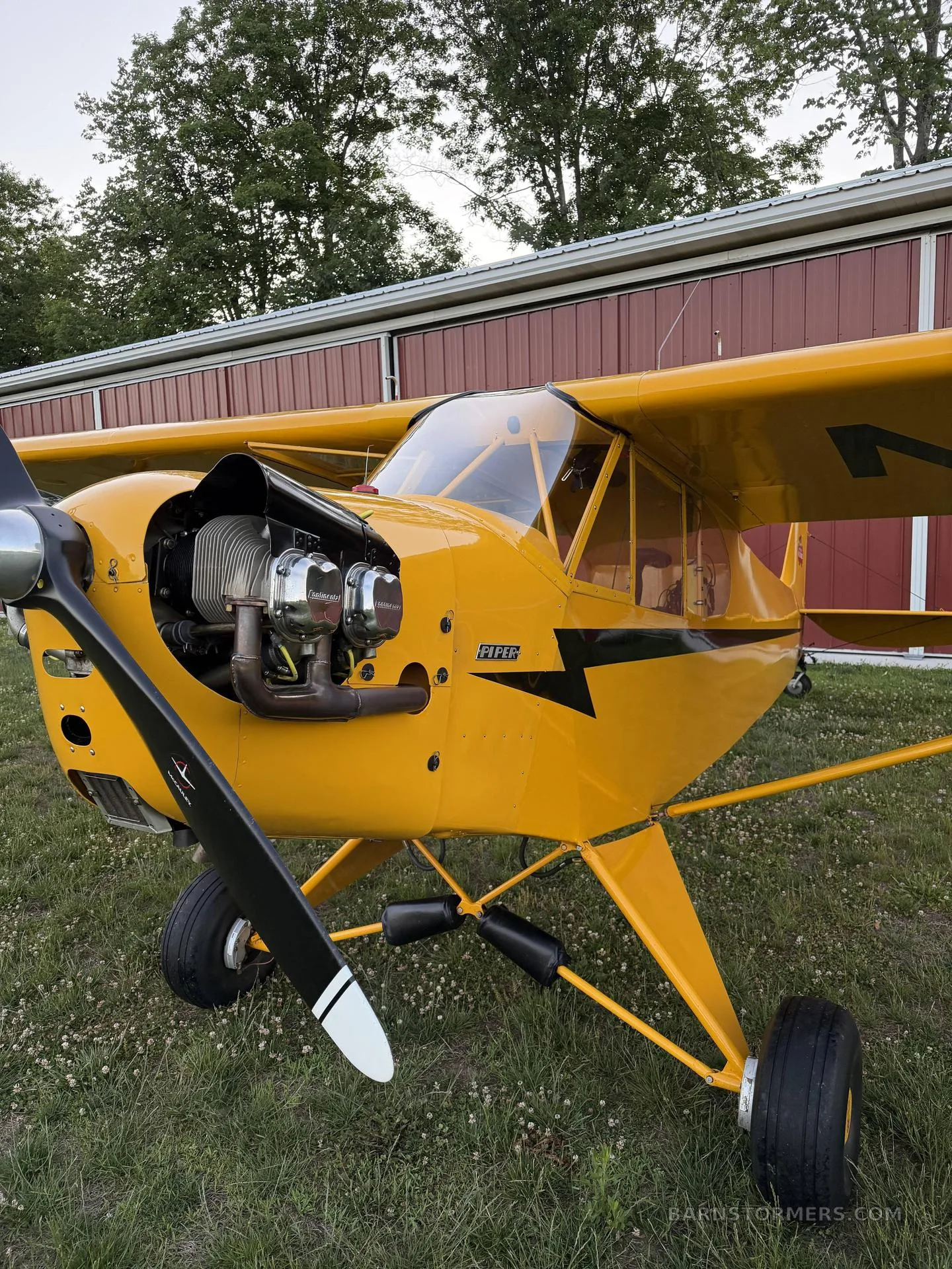A close-up front view of a yellow Piper Cub aircraft with its propeller and exposed engine, parked on grass.