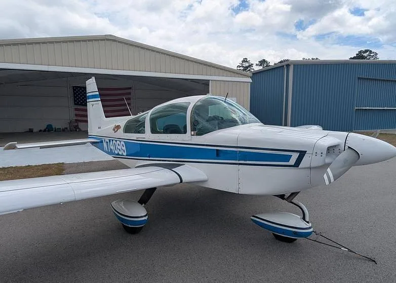 A front-right view of the white and blue Grumman AA-5 Traveler N74099 parked in front of an open hangar with an American flag inside.
