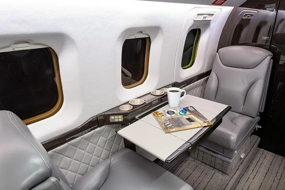 A close-up of the business jet's cabin interior, showing gray leather seats, a fold-out table with a coffee cup, and windows.
