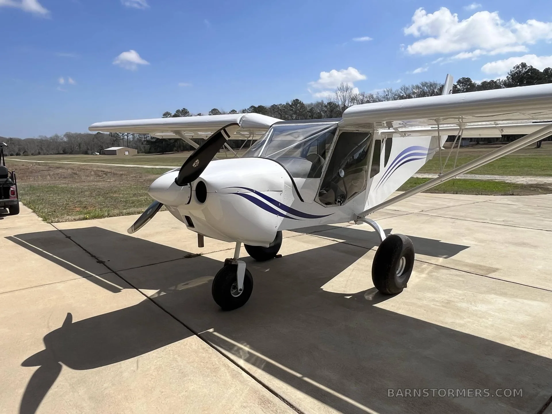 A white high-wing aircraft with blue stripes on the fuselage, parked on a concrete pad under a clear sky.